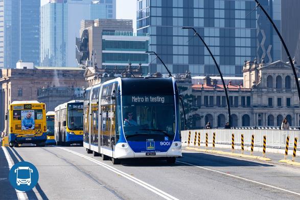 The image shows a modern city street with a metro bus in the foreground. The bus is blue and white with the words "Metro in testing" displayed on the front electronic sign. It is travelling along a multi-lane road with protective barriers on either side. Behind, several other buses are visible, with the closest being yellow. The background features a mix of historical and modern architecture, with tall office buildings and a historic structure with the text “Q. BANK OF NEW SOUTH WALES.” Additionally, there are people walking on the pavement adjacent to the bus route, and street lamps are uniquely angled above the road.