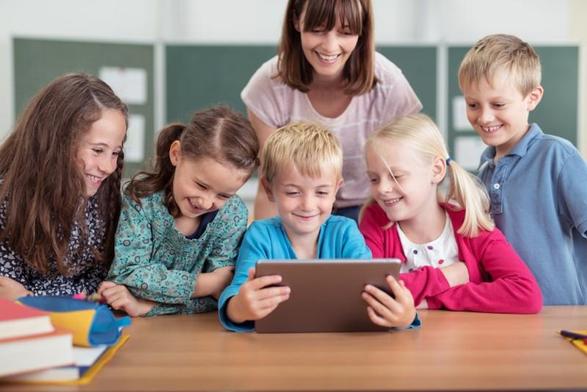 Fünf Grundschüler, die gemeinsam ein Tablet in der Hand halten und mit ihrer Lehrerin, die hinter ihnen steht, draufschauen.
