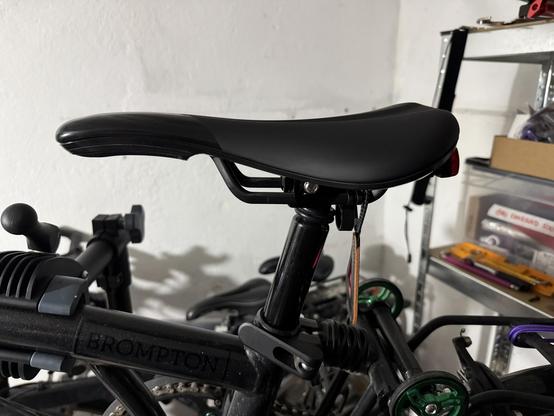 A close-up view of a bicycle seat, specifically from a Brompton folding bike, mounted on a black seat post. The background shows parts of other bicycles and tools.