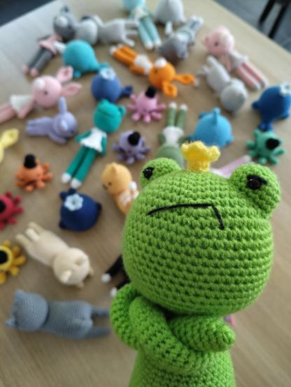 In the foreground a crocheted frog with his arms folded is looking grumpily at the viewer. In the background is a variety of crocheted animals spread out over a desk.