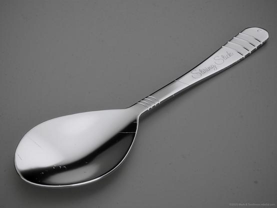 Silver coloured teaspoon with stiring stick engraved on the handle