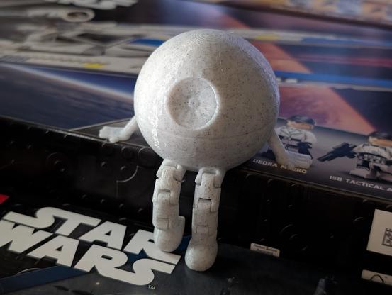 Photo of a 3D printed Death Star but with arms and flexible legs, so it appears mildly anthropomorphic. It is "sitting" on the edge of a Lego set box with its legs dangling. One foot is flat on the surface beneath it, the other is resting on its tiptoe.