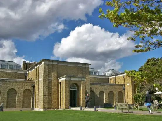 The image shows a building with a brick facade, surrounded by greenery and a blue sky with fluffy clouds. There are people walking nearby, and the area has benches and trees. The architectural style gives it a modern yet classic appearance.

Image Credits: Wikimedia / Poliphilo / CC0