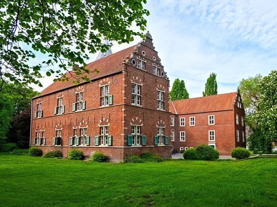 The Haneburg is a two-wing U-shaped residential castle made of red brick. It consists of a ground floor, 1st floor and an attic. The windows are small, some have green shutters and the building is surrounded by lawns and trees.
