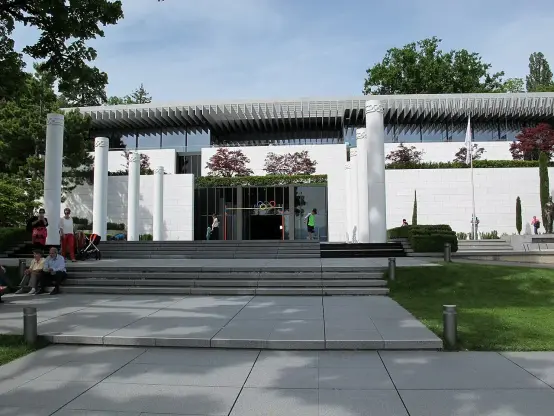 The image depicts a modern architectural building with a white exterior, featuring a series of columns and a wide staircase leading up to it. The entrance is framed by large glass doors, and there are people visible around the area, some sitting on the steps and others walking. The surrounding landscape includes greenery, such as trees and neatly trimmed bushes. The overall setting appears to be a well-maintained outdoor space, possibly part of a cultural or public institution.

Image Credits: Wikimedia / Sailko / CC BY-SA 4.0