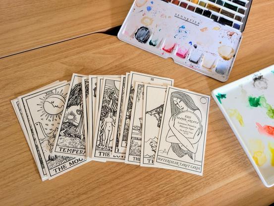 A set of Watercolor tarot cards spread on top of a table next to a watercolor palette. The cards have not being painted yet.