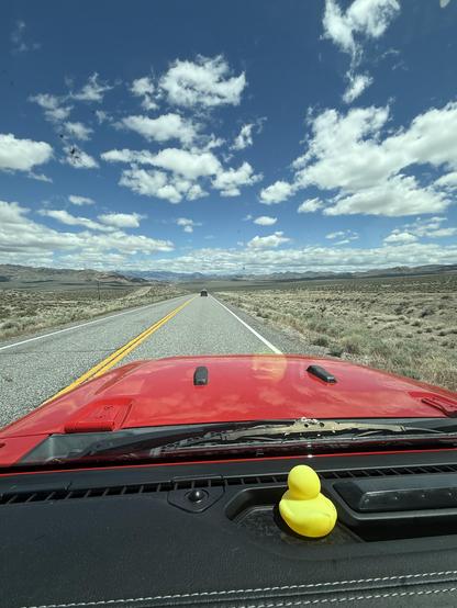 A red car is driving on a road with both sides of the road are sagebrush-covered field. Blue sky with patchy clouds. A yellow rubber ducky on the dash.