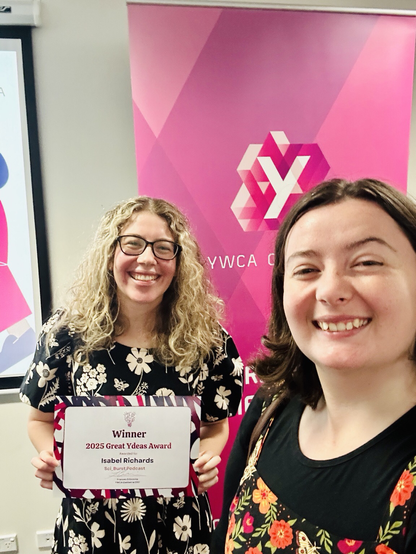 Two young women in floral dress standing in front of a pink YWCA banner. The one on the left is holding a grant certificate