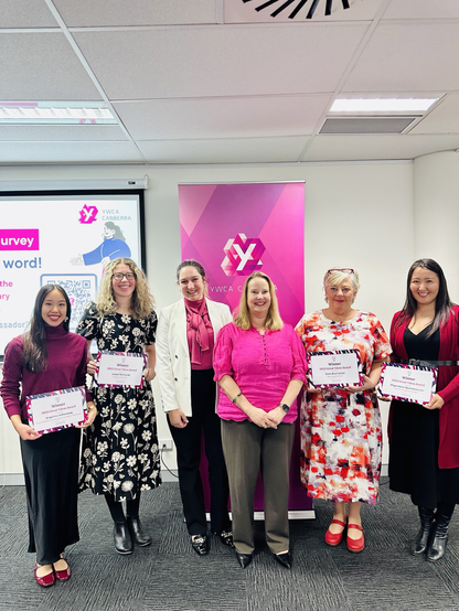 Six women (four of whom are holding grant certificates) standing in front of a pink YWCA banner