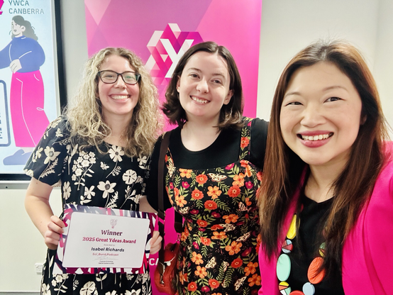 Three women wearing florals and pink in front of a pink YWCA banner. The one on the far left is holding a grant certificate