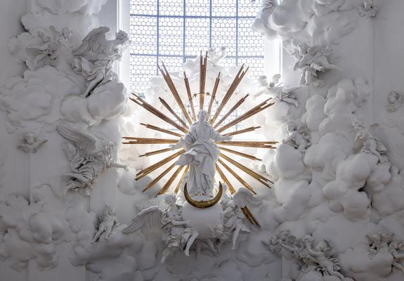 Baroque altar sculpture inside the Kollegienkirche in Salzburg, featuring a central figure of the Virgin Mary ascending amidst a burst of golden rays, surrounded by detailed white stucco clouds and numerous angels in dynamic poses, all illuminated by natural light from a large, grid-patterned window behind.