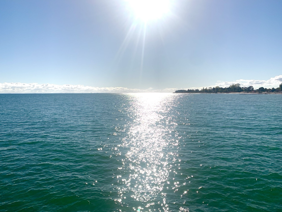 Die Ostsee im Gegenlicht der Sonne, die angescgnitten wurde am oberen Biksra d. Keine Wellen, sie Sonne spiegelt sich im blauen Wasser. Wenige Wolken am Horizont und etwas Küste rechts. oktober 2024