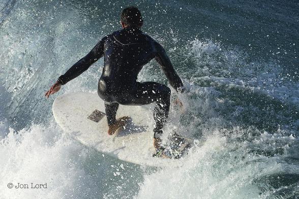 This Is A Colour Close-Up Water Sport Photo In Landscape Format. Above And To The Left Of Centre Is The Back Of A Male Surfer Wearing A Black Wetsuit. He Is Surfing On A White Surfboard Diagonally Across The Frame From Right To Left. The Surfer Is On The Crest Of The Wave With White Surf Below Him, Spray And Water Droplets All Around. The Early Morning Sun, Still Low, Is Illuminating The Front Of The Breaking Wave And The Surfer's Left Side. The Ocean Side Of The Wave Is In Shadow. Our Surfer Is Standing On His Toes As He Balances On His Board, He Is Crouched With His Knees Bent And Arms Slightly Outstretched. 
The Location: Hermosa Beach, California (2013).