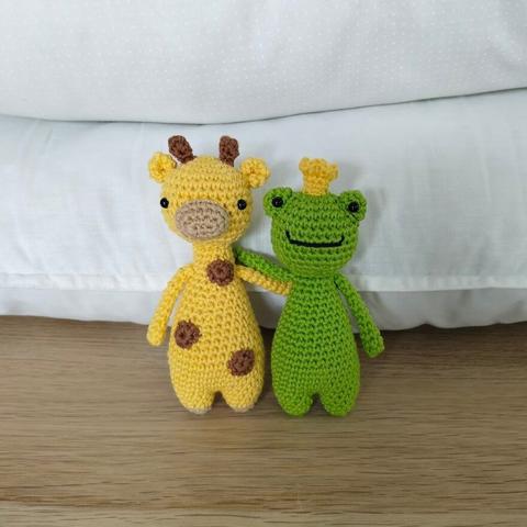 A small crocheted giraffe and frog with a yellow crown are standing next to each other, arms around each other's shoulders.