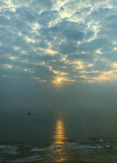 Photo of a beach in the early morning, taken from a high vantage point. The sun pierces through a dramatic cloud-filled sky and is reflected by the sea in a glittering column that stretches to the shore. Distant smog softens the horizon so that it’s hard to tell where the sea ends and the sky begins. Tiny human figures dot the sand and wade into the water to meet foam-edged waves rolling ashore. Tiny boats are scattered here and there on the sea.

I think this image conveys both how small we are in the vastness of our world and yet how destructive we can be to it. The people in the image are tiny as ants. And yet it’s people who create the smog that has made the horizon disappear, a beautiful and disturbing sight all at once. In more ways than one, our horizons, our possible futures, are disappearing today because of our shortsighted choices. And yet beyond the smog and the clouds the sun still shines, beyond our reach, a reminder that the great and terrible beauty of the universe will outlast us all. And I suppose I take some comfort in that.