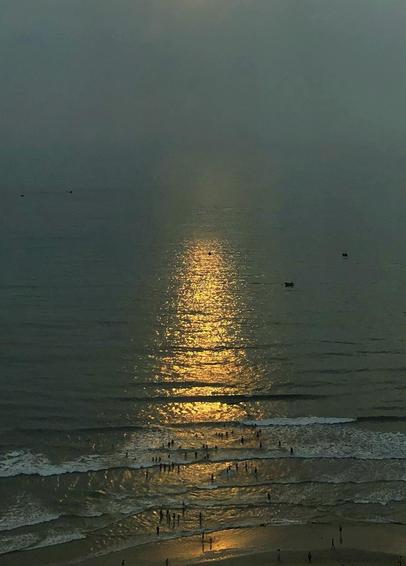 Cropped version of the previous photo,  focusing on the column of sun glitter on the sea extending to the people on the shore, illuminated by the glow as they stand amid the waves.