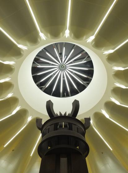 The image features a unique architectural design, showcasing a circular ceiling with a radial pattern of lights. At the center, there is a circular opening that draws the eye upward, while the lower portion features a large, stylized structure that resembles a pedestal or sculpture. The color palette is predominantly soft and warm, enhancing the modern aesthetic of the space.