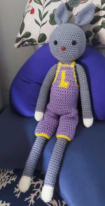 Picture of a crochet long-legged bunny amigurumi. It's gray, with a round head, pointed ears and long legs and arms. The paws part of the arms and legs are white. It has black safety eyes and a little brown nose. It's wearing a pair of crochet overalls in purple and yellow. The amigurumi is sitting on a pile of blue pillows.