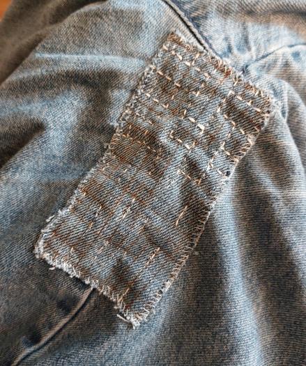 Das Bild zeigt einen Flicken auf einer Jeans. Man erkennt vorgezeichnete Linien und einige unzusammenhängende gestickte Linien. Das Ganze ist auch noch krumpelig verzogen.