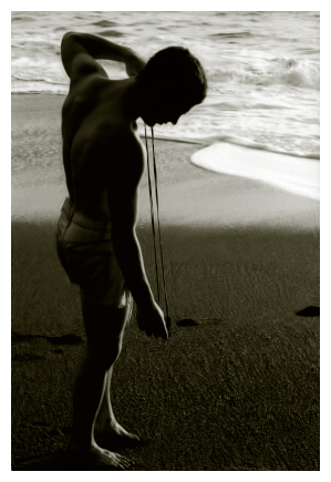 monochrome. late afternoon on a pacific beach. a mid-20s man in white, cut-off denim shorts stands on the beach, at the water's edge in silhouette, left arm raised, and right hand holding a DIY rawhide slingshot. the sand is black and small waves recede after soaking it.