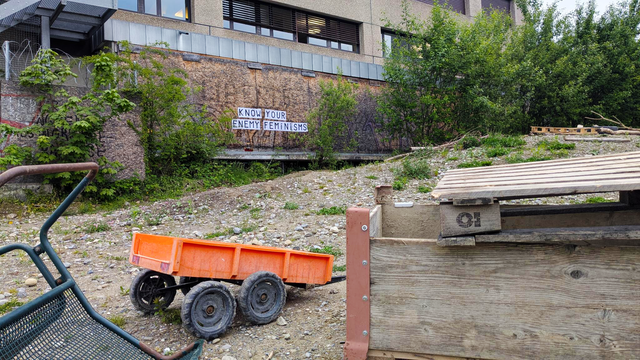 In the foreground, an upturned chair, an orange toy truck and a wooden crate on a gravel lot. In the background, a house with a wooden wall and trees/bushes. On the wooden wall a feminist collage that reads: KNOW YOUR ENEMY FEMINISMS
