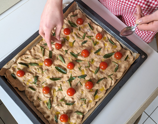Eine Person bereitet gerade frische Foccacia zu, belegt mit frischen Kräutern (Salbei und Rosmarin) und roten Tomaten. Foto von Karen Kaspar.