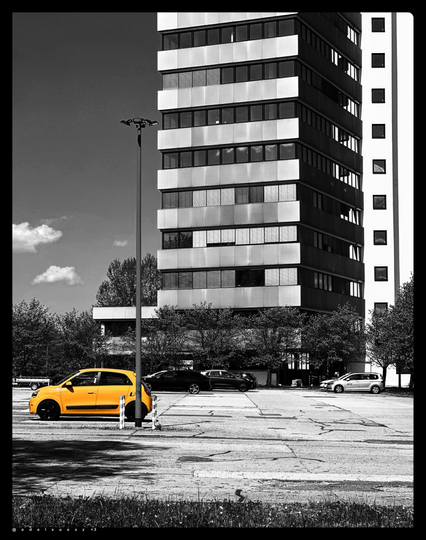 Schwarzweißfotografie mit einem Farbakzent: großer betonierter Parkplatz vor einem Hochhaus, mehrere parkende Autos, ein Fahrzeug im Vordergrund nur in leuchtend gelber Farbe, ein Kleinwagen Renault Twingo