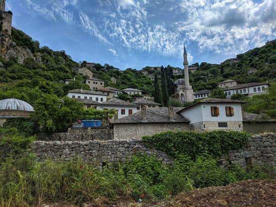 A view of Počitelj, Bosnia and Herzegovina, showcasing a historic village nestled against a rocky hillside. The foreground features green vegetation, leading to a stone wall that borders the village. Traditional white-walled houses with red-tiled roofs are visible, interspersed with stone buildings and a van. A slender minaret rises in the background, adding to the architectural tapestry. The hillside is lush with greenery, and the sky is a mix of blue and fluffy white clouds.
