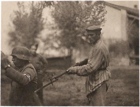 Ein Mann in gestreifter Kleidung, der offensichtlich aus KZ Haft befreit wurde und mit einer Waffe auf einen Nazi-Soldaten zielt, welcher die Hände hoch hält. Im Hintergrund ist ein alliierter Soldat zu sehen, der die Szene beobachtet.