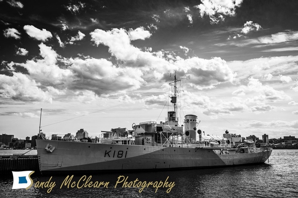 Small historic warship alongside under a partly cloudy sky against a city skyline.