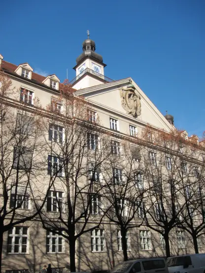 The image shows the exterior of a large building with a clock tower on top. The architecture features a blend of classical elements, including a decorative façade with a central emblem, likely representing an institution or organization. The surrounding area has bare trees, indicating it might be early spring or late autumn, and the sky is blue, suggesting clear weather. The building appears to be well-maintained and could be associated with educational or civic purposes.

Image Credits: Wikimedia / Theresia-Gerhardinger-Gymnasium am Anger / Public domain