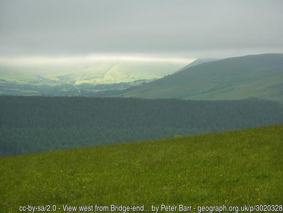 A series of dark hills below low cloud. In the remote distance a patch of sunlight floods over a green slope