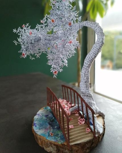 Création unique. Grande sculpture d'un cerisier en papier blanc. L'arbre est recouvert de part et d'autres de mots et de phrases imprimées en noir. Au pied de l'arbre, on voit un pont fait en carton, qui passe eu dessus d'un point d'eau, et qui est à moitié recouvert de fleurs de cerisier roses. Quelques fleurs sont encore accrochées dans l'arbre. La sculpture repose sur une bûche de bois coupée assez fine, qui fait office de socle.