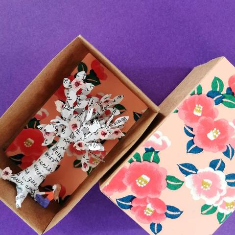Un arbre en papier blanc dans une petite boîte qui peut se refermer (comme une grande boîte d'allumette). L'arbre est recouvert de texte en français et japonais, imprimé en noir (on y aperçoit notamment le mot "cerisier"). On y voit également quelques fleurs de cerisiers roses au niveau des feuilles. Une femme vue de côté se tient sous l'arbre en tailleur, face à ce dernier. Le fond et le dessus de la boîte est rose clair, avec des motifs de fleurs de cerisiers roses et de feuilles vertes.
