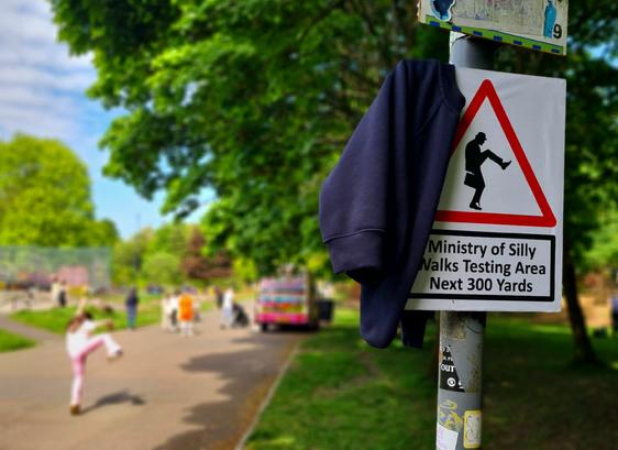 A sign of a man walking in a strange way in a red triangle. Beneath it are the words Ministry of Silly Walks Testing Area Next 300 Yards. To the left of this is a young child walking in a strange way.