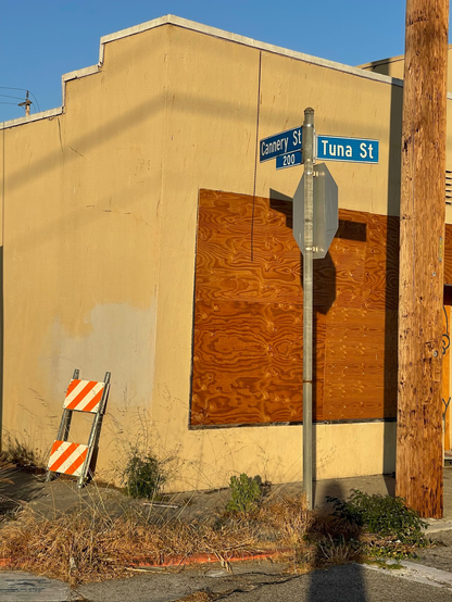 A one-story, boarded up building in bright sunlight, at the intersection of Cannery and Tuna Streets, Terminal Island
