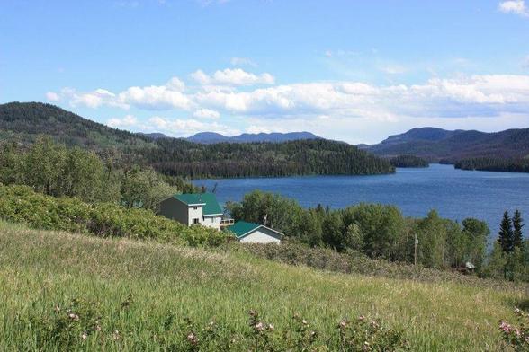 A blue lake nestles between grassy meadows and the dark tree covered hills in the distance.