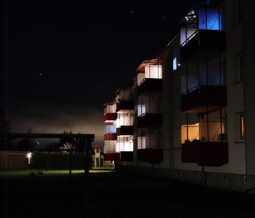This image shows an apartment building at night. The building has several balconies, some of which are lit up with different colors of lighting. The sky is dark with visible stars, and there is some ambient lighting in the surrounding area.