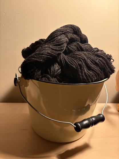 A yellow enameled metal bucket, resting on a light-coloured wooden bench. The bucket is piled high with naturally coloured black (well, really dark brown) yarn.