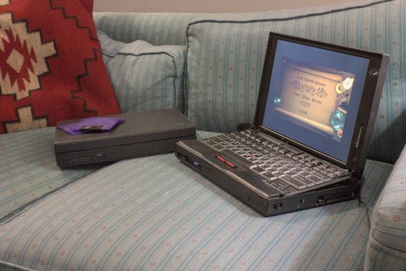 Thinkpad 760EL opened up next to a closed 760CD. The 760EL has a 760CD badge on it because the keyboard has been transplanted. The screen is displaying the first menu in Daggerfall. This screen is not severely damaged/flawed like the one that came with the 760EL was. That's now on the 760CD which is relegated for parts use. Both laptops are on a couch, there are some translucent purple floppy disks stacked on the closed lid of the 760CD.