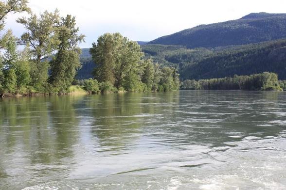A ripple in the water of a smooth river that flows between rocky and tree lined banks toward the rounded hills in mid and far distance.