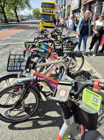 A row of shared bicycles completely filling a section of bike stands on a city street. In the background, people walk on the sidewalk and a yellow double-decker bus is visible.