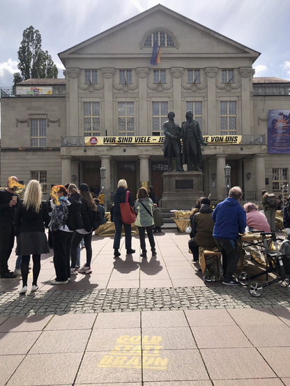 Theaterplatz in Weimar mit Blick auf die Fensterfassade des Nationaltheaters, davor haben sich viele Menschen versammelt um den 80. Jahrestag des Kriegsendes zu gedenken; das Denkmal mit Goethe und Schiller ragt über ihnen auf und an der Fassade des DNT ist ein goldenes Spruchband mit dem Text: „Wir sind Viele“, auf dem Boden des Platzes ist in goldener Farbe „Gold statt Braun“ gesprüht.