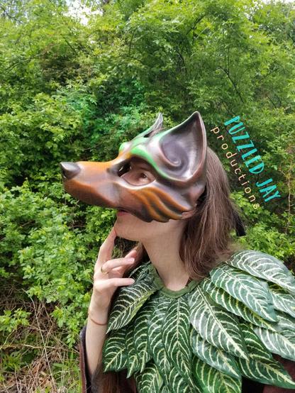 people wearing a brown and green wolf mask peer around trees