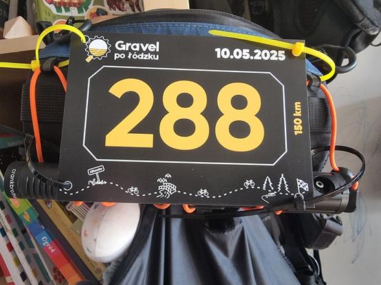 Background: book shelf and backpacks. Main plan in the middle: black thick paper with yellow number 288 surrounded by white border. At the bottom there's simple white ornament through whole width of the paper with trees, bikes and bridges. On the right horizontally there's is yellow text 150km. Top left corner: logo and text Gravel po łódzku. Top right corner: date in white - 10.05.2025

Whole piece of paper is tied with two yellow zip ties in top corners and two black zip ties in the bottom corners to the barely visible handlebar bag. Underneath there's a small black air pump
