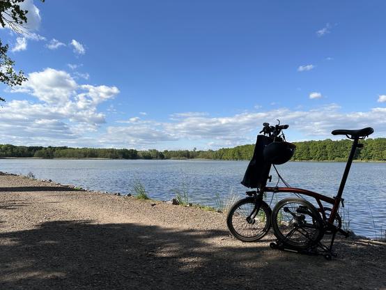 Photo of my semi-folded Brompton on a gravel road next to large body of water with tree line in the background.