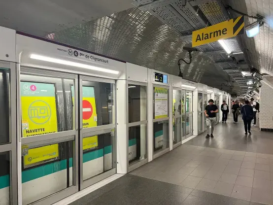 The image shows a subway station, likely part of a metro system. There's signage indicating directions, with specific references to "Pte de Clignancourt" and "Alarme." The station features train doors that are closed, and there are people walking along the platform. The overall environment appears modern and well-maintained. The colors and graphics suggest information related to transit services.

Image Credits: Wikimedia / Chabe01 / CC BY-SA 4.0