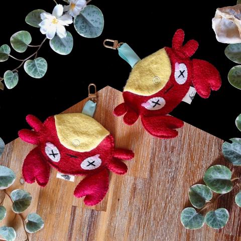 Two crab pouches side by side. They’re hanging upside down by a satin ribbon that links them to a small lobster claw. Their bellies are yellow and they have X-s in their eyes, indicating that they’re playing dead.
