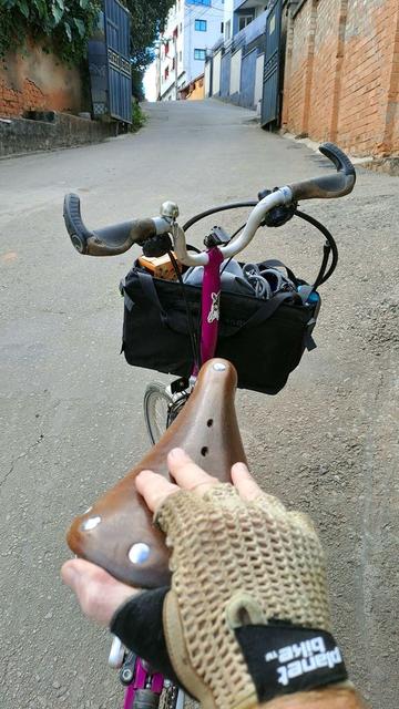 A person wearing a bike glove pushes a pink bicycle up a steep hill in a residential neighborhood in Antananarivo, Madagascar. The photo is taken from their perspective, showing their hand on a brown leather saddle and the bicycle's handlebars, which are equipped with a front bag and a “Doozy” sticker on the stem, referencing a Great Dane influencer