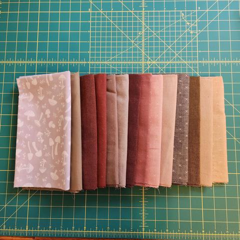 14 pieces of folded half yard cotton. They are a collection of warm autumn/earthy tones. Some are plain and some have small texturing in the form of hatches. One piece is grey with several white mushrooms on it.